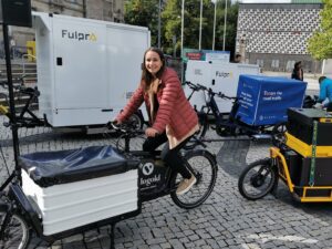 Jana Dornieden von der Automotive Agentur sitzt auf einem E-Lastenrad mit einem weißen Container im vorderen Bereich, der mit einer schwarzen Plane abgedeckt ist.