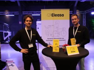 Niklas Nötzel, Head of Sales, Vertriebsmanagement der Firma Eleasa mit einem Kollegen an einem Stehtisch mit Rollup im Hintergrund.