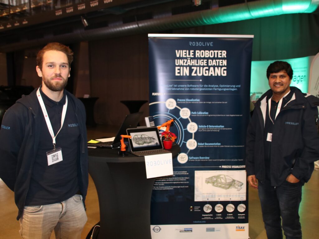 Arne Brökers von der Firma Robolive und ein Kollege rechts und links von einem Rollup und einem Stehtisch mit Miniroboterarmen und einem Laptop, der das Überwachungssystem für Roboter zeigt