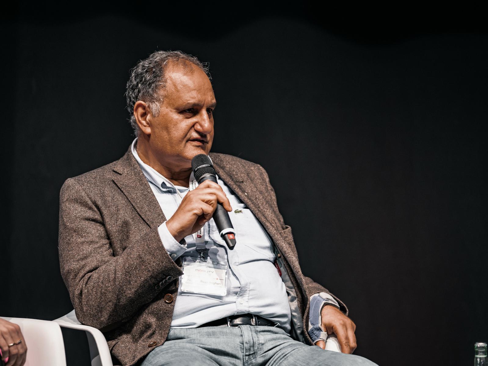 Prof. Reza Asghari von der TU Braunschweig sitzt auf der Bühne in einem weißen Stuhl und spricht in ein Mikrophon, das er in der Hand vor seinen Mund hält.