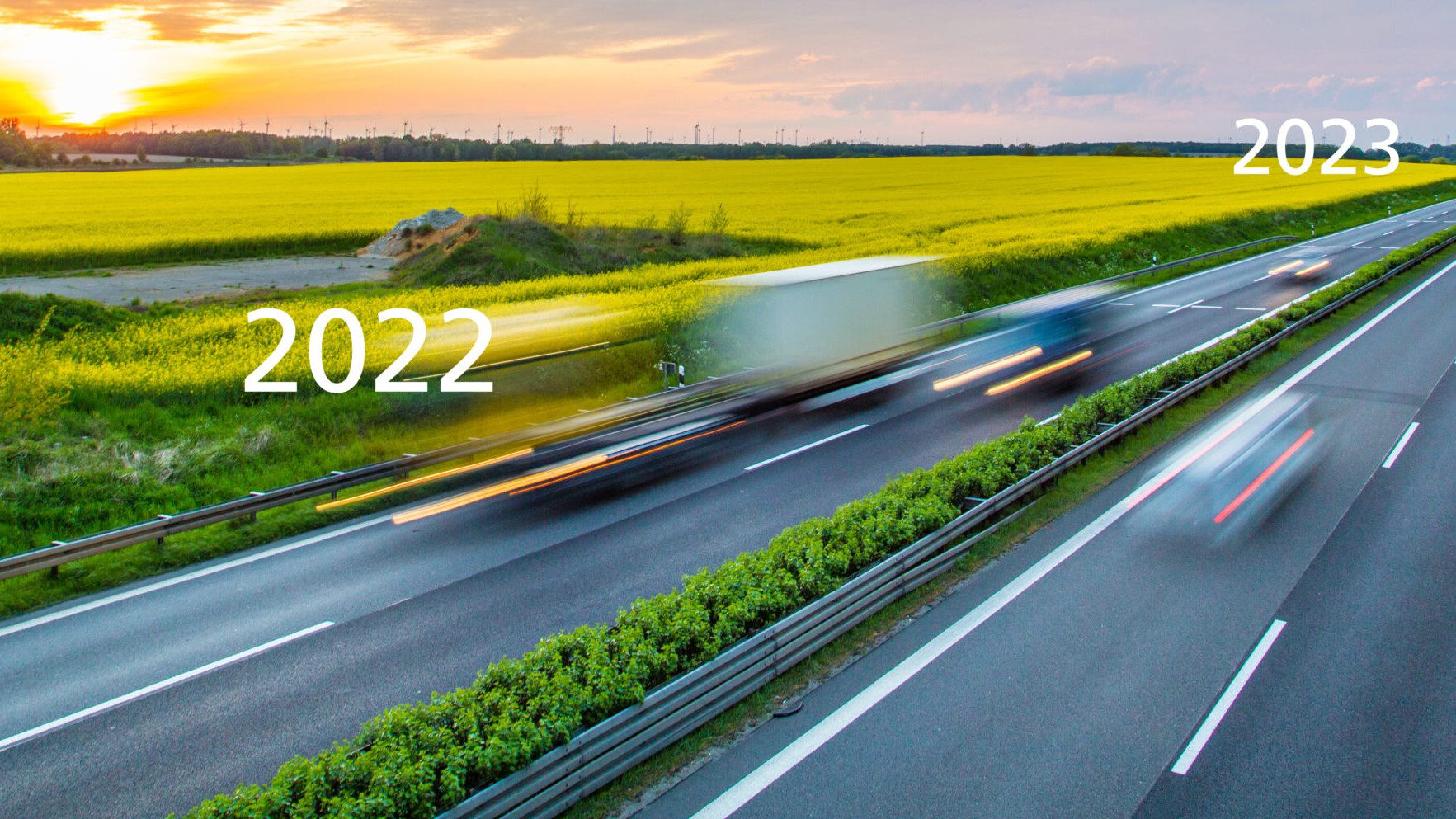 Langzeitaufnahme von fahrenden Autos und Lastern auf einer Autobahn, im Hintergrund ein Rapsfeld und Windkraftanlagen im Sonnenuntergang.