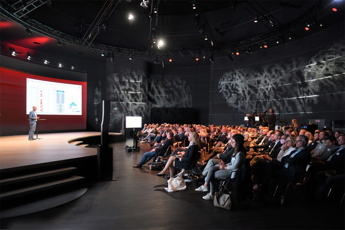 Blick in eine Veranstaltungshalle: Ein Redner auf de Bühne mit Projektion hinter sich. Gefüllte Zuschauerränge.