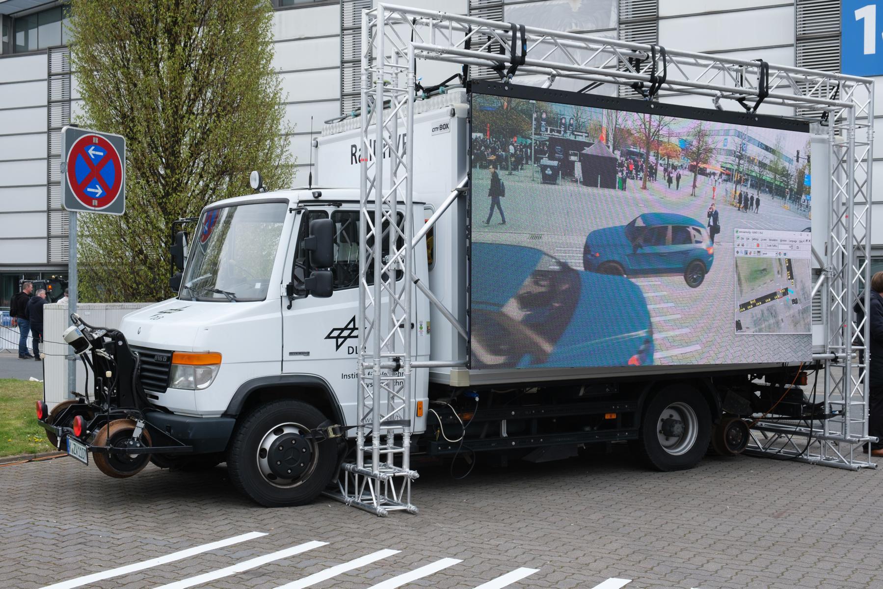 Eine Lastwagen mit einer großen Leinwand, auf der die Umgebung sowie virtuelle Autos zu sehen sind. Davor ein Zebrastreifen.