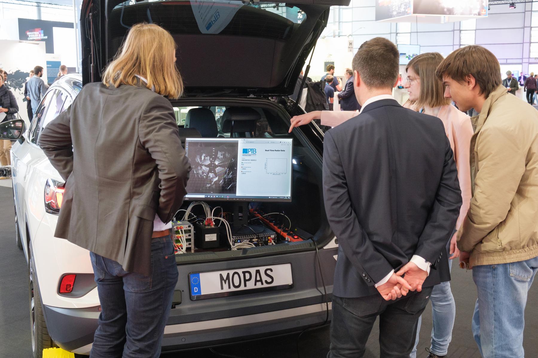 Eine Gruppe Menschen blickt auf ein Display im geöffneten Kofferraum eines Autos.